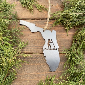 May include: A silver metal ornament shaped like the state of Florida with a silhouette of a person paddleboarding inside. The ornament is hanging from a twine string.