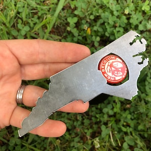May include: A metal bottle opener shaped like the state of Virginia. The opener has a hole in the shape of a bottle cap, with a red and white bottle cap inside the hole. The bottle cap has the text "BELGIUM NEW BREWING" on it.