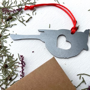 May include: A grey metal ornament shaped like a state, featuring a heart cut-out, and a red string for hanging. The ornament is set against a backdrop of greenery and a brown envelope, creating a rustic feel.