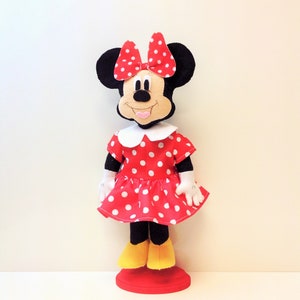 May include: A red and white polka dot dress with a white collar and a red bow on top of the head. The doll has black ears and a smiling face. The doll is standing on a red base.