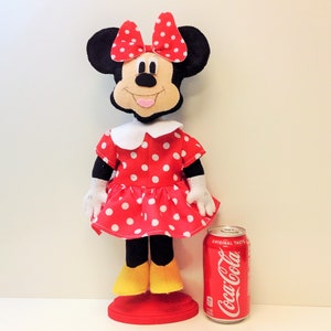 May include: A red and white polka dot dress with a white collar and a black bow. The dress is on a doll with black ears and a black face. The doll is standing on a red base. A can of Coca-Cola is in the background.