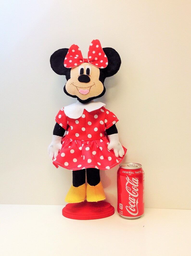 May include: A red and white polka dot dress with a white collar and a black bow. The dress is on a doll with black ears and a black face. The doll is standing on a red base. A can of Coca-Cola is in the background.