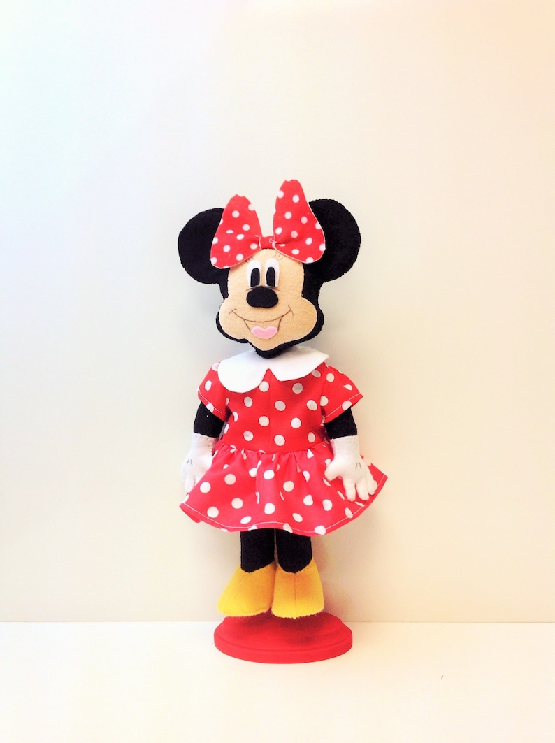 May include: A red and white polka dot dress with a white collar and a red bow on top of the head. The doll has black ears and a smiling face. The doll is standing on a red base.