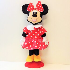May include: A red and white polka dot dress with a white collar and a black bow. The dress is on a doll with black ears and a black face. The doll is standing on a red base.