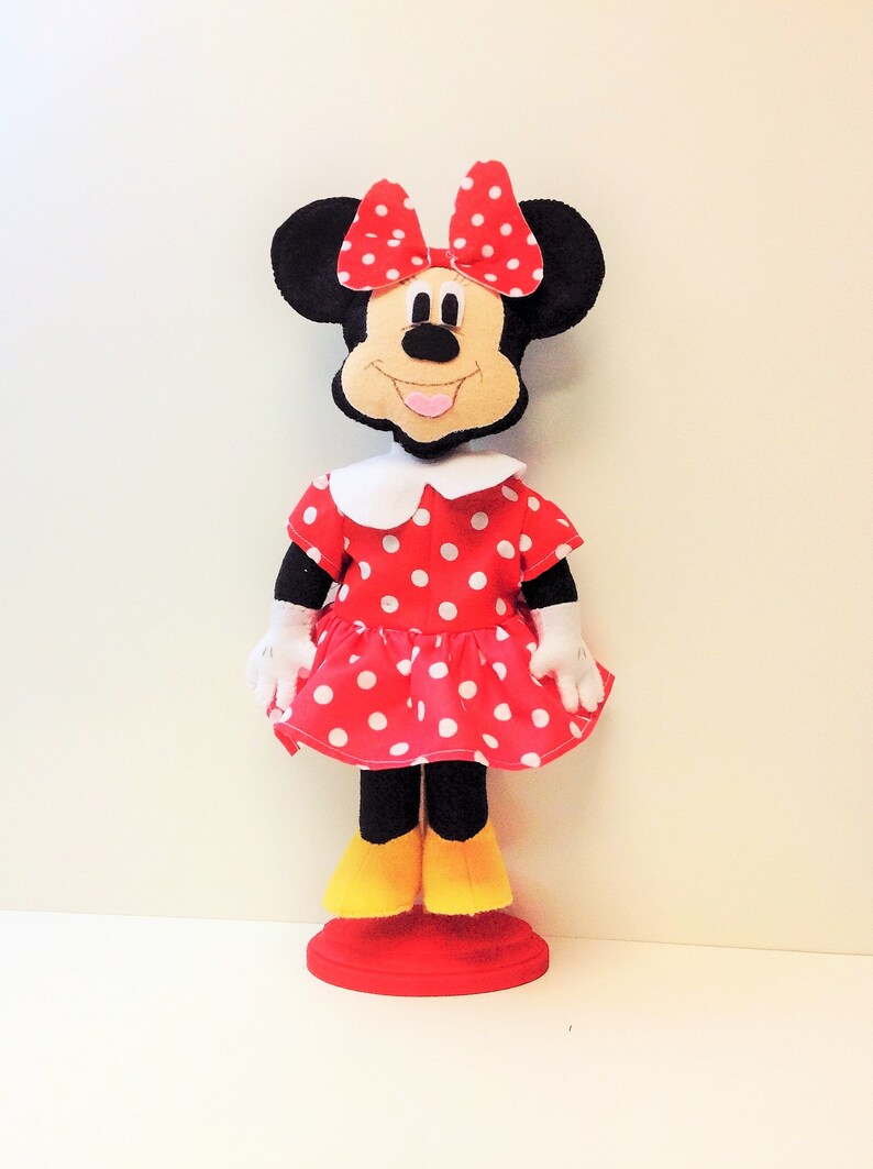 May include: A red and white polka dot dress with a white collar and a black bow. The dress is on a doll with black ears and a black face. The doll is standing on a red base.