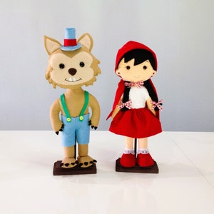 May include: A brown wolf wearing a blue and green outfit and a black top hat stands next to a red-hooded girl doll with black hair and a white dress. Both dolls are on wooden stands.
