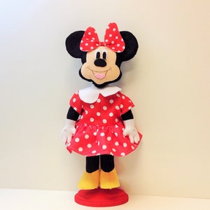 May include: A handmade plush toy of Minnie Mouse wearing a red dress with white polka dots and a red bow. The toy has black ears and a black nose. The toy is standing on a red base.