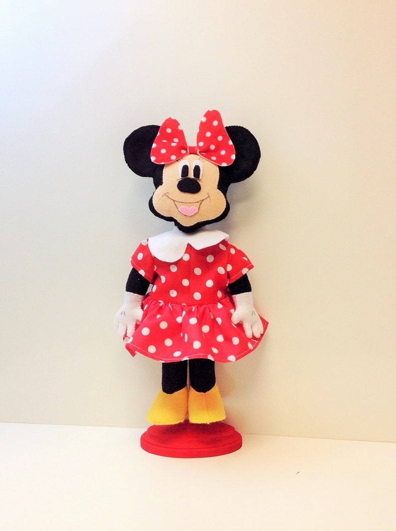 May include: A handmade plush toy of Minnie Mouse wearing a red dress with white polka dots and a red bow. The toy has black ears and a black nose. The toy is standing on a red base.