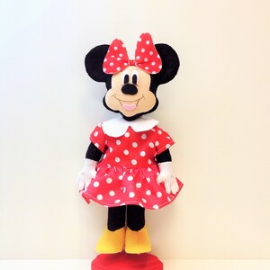 May include: A red and white polka dot dress with a white collar and a red bow on top of the head. The doll has black ears and a black nose. The doll is standing on a red base.