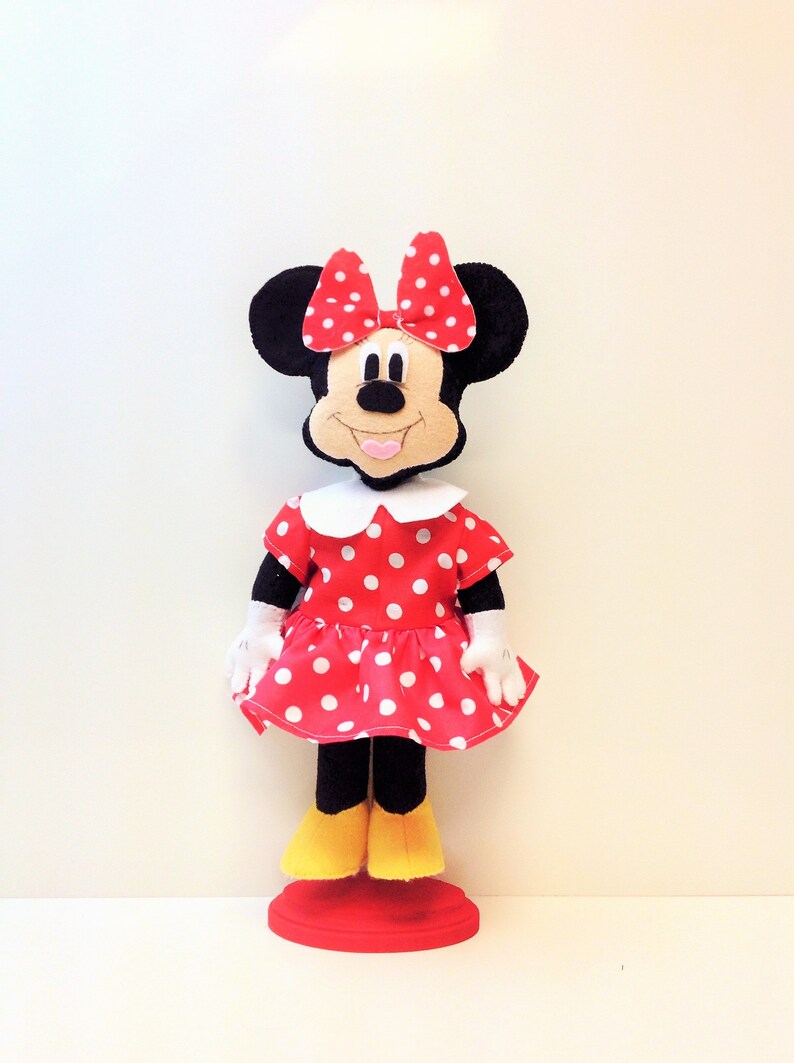 May include: A red and white polka dot dress with a white collar and a red bow on top of the head. The doll has black ears and a black nose. The doll is standing on a red base.