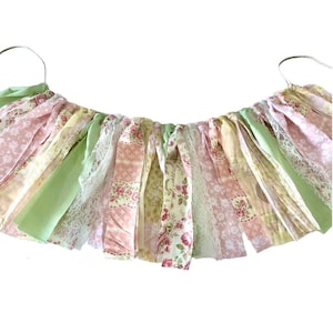 May include: A green, pink, and yellow fabric garland with floral patterns and lace trim. The garland is strung on a white cord and is perfect for adding a touch of whimsy to any party or event.