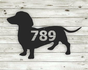May include: Black silhouette of a dachshund with the number 789 cut out of the body. The silhouette is on a white wood background.