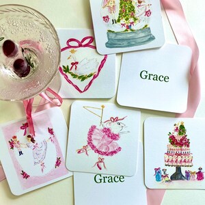 May include: A collection of square coasters with watercolor illustrations. Designs include a snow globe, a swan, a ballerina mouse, a tiered cake, and a ballerina. Some coasters have the name "Grace" printed on them. A glass of liquid with cherries is in the upper left.