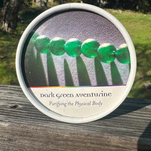 May include: A round container with a white rim, featuring a string of dark green aventurine beads. The text "dark green aventurine" and "Purifying the Physical Body" is printed on the container. The background is a gradient of colors.