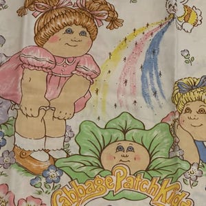 Pode incluir: Um lençol branco com personagens Cabbage Patch Kids. O design inclui uma menina com um vestido rosa, um rosto em forma de repolho e um arco-íris. As palavras "Cabbage Patch Kids" estão em uma faixa amarela.