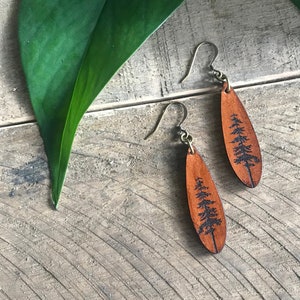 Può includere: Un paio di orecchini in pelle marrone con un design ad albero. Gli orecchini sono appesi a una superficie di legno.