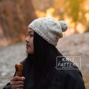 May include: A white and grey speckled knit beanie with a fluffy pom pom on top. The beanie is being worn by a person with long black hair. The text "KNIT PATTERN @VONNIEANDCO" is visible in the lower right corner of the image.