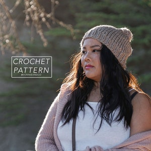 以下が含まれることがあります： 女性が、ポンポンが付いたベージュのクロシェのビーニーをかぶっています。ビーニーは、テクスチャのあるステッチで作られています。女性は、白いタンクトップとピンクのカーディガンを着ています。背景はぼやけていて、ピントが合っていません。画像には、「CROCHET PATTERN @VONNIEANDCO」というテキストが表示されています。