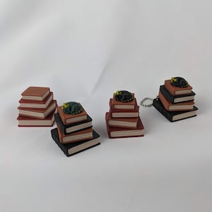 Pode incluir: Pequenas pilhas de livros em tons de castanho e preto, algumas com uma figura de gato a dormir em cima. Uma pilha está presa a um porta-chaves prateado. Os livros têm páginas avermelhadas e estão dispostos em pequenas pilhas decorativas.