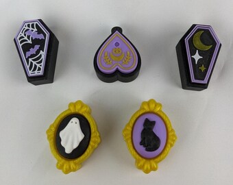 May include: A collection of Halloween-themed decorative items. The set includes two coffin-shaped pieces with bat and moon designs, a heart-shaped piece, and two oval-framed pieces with a ghost and a black cat. The items are black, purple, gold, and white.