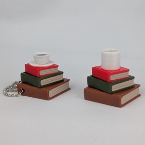 Puede incluir: Miniaturas de pilas de libros con una taza y platillo pequeños, y un cilindro blanco. Las pilas son marrones, verdes y rojas, con un borde blanco. Una pila tiene una cadena plateada.