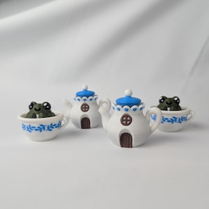 Könnte beinhalten: Eine skurrile Sammlung von Miniatur-Keramik-Teekannen und Tassen. Die weißen Teekannen haben blaue Akzente und braune, türförmige Details. In jeder Tasse und Teekanne sitzt eine kleine grüne Froschfigur. Die Tassen und Teekannen sind mit blauen Blumenmustern verziert.