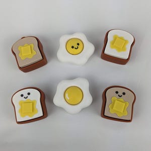 以下が含まれることがあります： 食べ物をテーマにした6つの装飾チャーム。黄色の卵黄と白い卵白の目玉焼き2つと、黄色のバター正方形のトースト4切れ。トーストは茶色の地殻とタンパンです。トーストスライスの中には笑顔のものもあります。