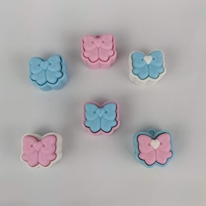 Puede incluir: Seis colgantes con forma de lazo en tonos pastel. Cada colgante presenta un diseño de lazo con un corazón en el centro. Los colores incluyen azul claro, rosa y blanco, con un contorno contrastante.