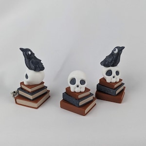 Könnte beinhalten: Drei Miniaturskulpturen mit einem schwarzen und weißen Raben, der auf einem weißen Schädel sitzt, der auf einem Stapel brauner und grauer Bücher ruht. Der Hintergrund ist schlicht weiß. Dekorative Objekte.