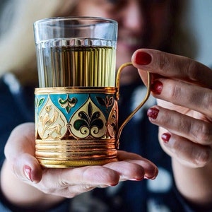 Könnte beinhalten: Ein goldfarbener Metall-Tee-Cozy mit blauen und grünen Emaildetails und filigranen Blumenmustern. Der Cozy hält eine Glas Tasse mit einer hellfarbigen Flüssigkeit.