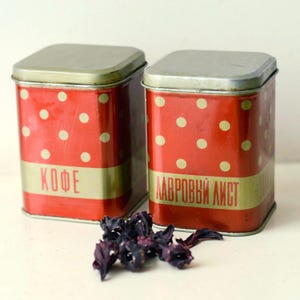 Puede incluir: Dos latas de almacenamiento de metal rojo con lunares dorados y texto en idioma extranjero. Las latas tienen una banda dorada en el medio y tapas plateadas. Flores moradas secas están delante de las latas.