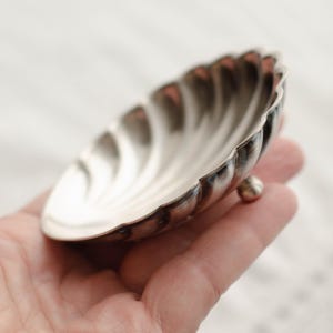 Könnte beinhalten: Eine kleine, silberfarbene, muschelförmige Schale mit gewelltem Rand und drei kleinen Kugelfüßen. Die Schale hat eine polierte, reflektierende Oberfläche. Die Schale wird in einer Hand gehalten.