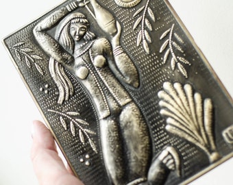 May include: A rectangular, bronze-toned metal relief sculpture featuring a stylized female figure holding a bottle. The artwork includes sun and leaf motifs, with a textured background and a decorative border. The sculpture is held in a hand.