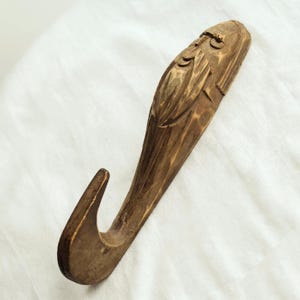 May include: A hand-carved wooden wall hook featuring the sculpted face of a bearded figure. The natural brown wood displays visible chisel marks and a smooth finish, with the hook extending from the lower portion of the elongated carved design.