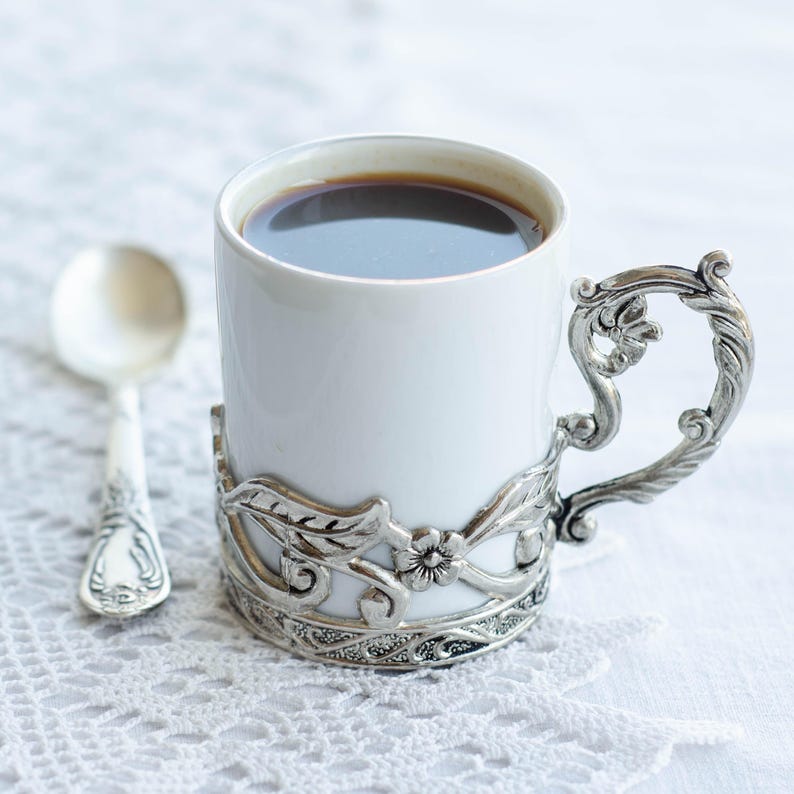 May include: A white ceramic coffee mug filled with dark liquid, held in an ornate silver holder with floral and leaf designs. A matching silver spoon sits beside the mug on a white lace surface. The mug's handle is also silver.