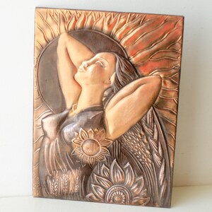 May include: A copper-toned metal wall sculpture depicting a woman with long hair, surrounded by sunflowers and wheat. The woman is depicted with her arms raised above her head, and her face is turned upwards.