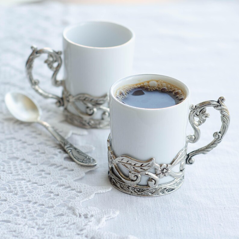 May include: Two white ceramic coffee mugs with decorative silver-coloured metal handles and bases, one filled with dark coffee. A silver spoon sits beside the mugs on a white lace tablecloth. The handles and bases have a floral design.