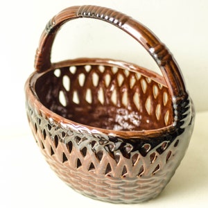 Può includere: Un cestino in ceramica marrone con manico. Il cestino presenta un motivo intrecciato con intagli a forma di diamante. Il manico è di un marrone più scuro e ha un aspetto strutturato e intrecciato. Il cestino è smaltato e ha una finitura lucida.