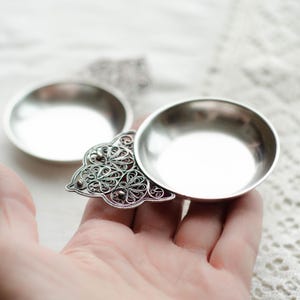 May include: A small, silver metal dish with a decorative filigree design. The dish has a round bowl shape and is connected to a detailed, openwork filigree element. The filigree features intricate patterns and small, rounded accents.