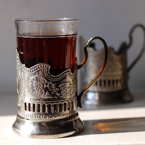 May include: A glass of tea in an ornate silver cup holder. The cup holder features a detailed floral design and a handle. Another cup holder is slightly out of focus in the background. The tea is a deep amber color.