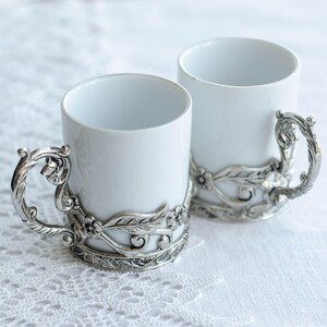 May include: Two white ceramic mugs with decorative silver-coloured handles and bands. The handles have a twisted design, and the bands feature floral and leaf motifs. The mugs are set on a white lace tablecloth.