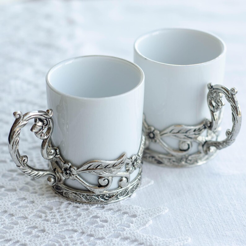 May include: Two white ceramic mugs with decorative silver-coloured handles and bands. The handles have a twisted design, and the bands feature floral and leaf motifs. The mugs are set on a white lace tablecloth.