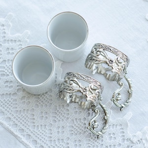May include: Three white ceramic cups with gold rims and two ornate silver napkin rings with floral and scroll designs are arranged on a white lace tablecloth. The napkin rings have a handle-like design.