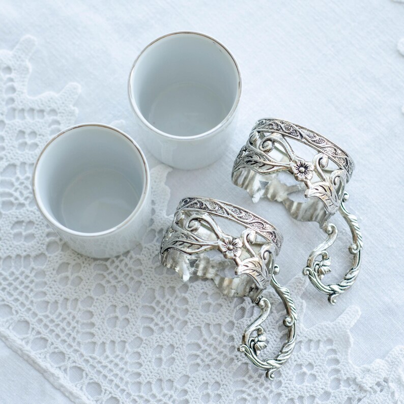 May include: Three white ceramic cups with gold rims and two ornate silver napkin rings with floral and scroll designs are arranged on a white lace tablecloth. The napkin rings have a handle-like design.