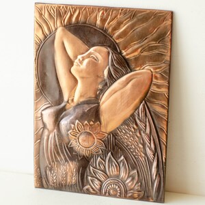 May include: A copper wall sculpture depicting a woman with her arms raised, surrounded by sunflowers and wheat. The sculpture has a textured, hammered finish.