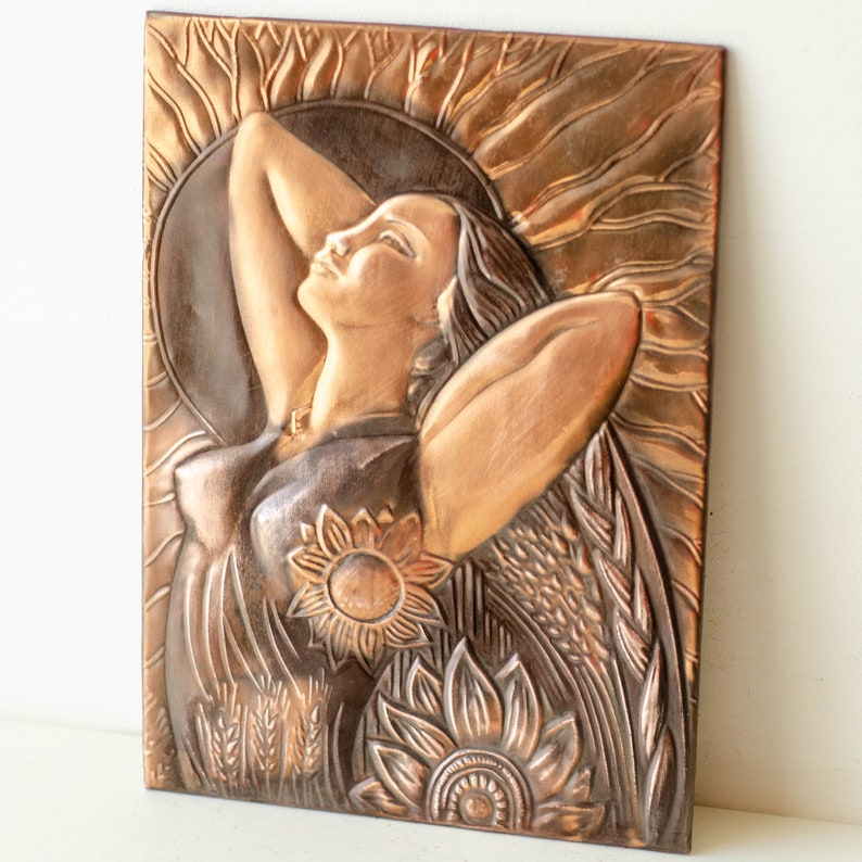 May include: A copper wall sculpture depicting a woman with her arms raised, surrounded by sunflowers and wheat. The sculpture has a textured, hammered finish.