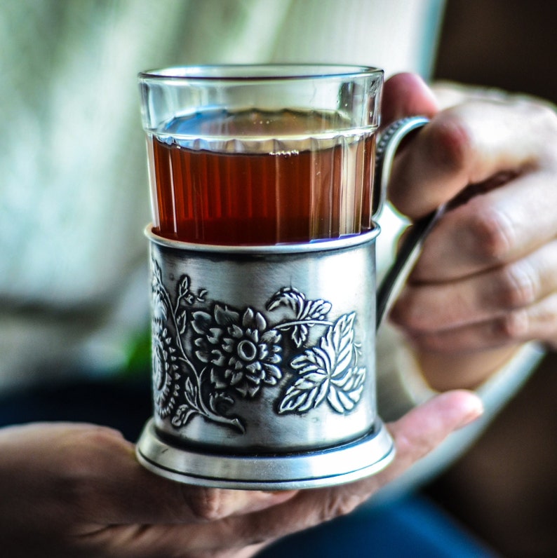 Op de afbeelding: Een theeglas van helder glas in een decoratieve zilverkleurige metalen houder. De metalen houder is voorzien van een reli&euml;f met een bloemmotief met gedetailleerde bladeren en bloemblaadjes. Het glas bevat een donker amberkleurige vloeistof.