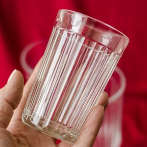 Puede incluir: Un vaso de vidrio transparente con líneas verticales, sostenido en una mano sobre un fondo rojo.
