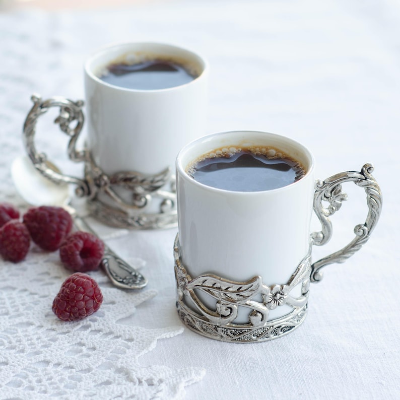 May include: Two white ceramic mugs filled with dark coffee, each with ornate silver-coloured metal handles and decorative bases. Fresh red raspberries and a small spoon are arranged nearby on a white lace tablecloth, creating a cosy, elegant setting.