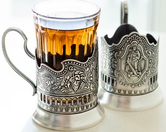 May include: A clear glass tea cup held within an ornate silver-toned metal holder featuring intricate floral relief patterns and a curved handle, accompanied by a matching empty silver-toned metal cup holder with a central figure relief design.